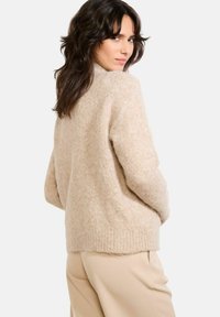 Beige adīta džemperis ar mīkstu, pūkainu tekstūru, apaļu dekoltē un ribotu apakšmalu. Dizains ietver brīvu piegriezumu un nokritušus plecus.