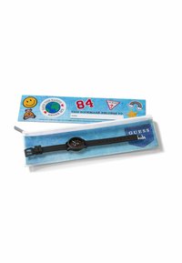 Orologio da polso nero per bambini con numeri rossi, mostrato su una custodia con motivo in denim e un segnalibro colorato con adesivi e etichetta "Guess kids".