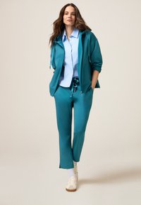 Türkisfarbiges Sportoutfit mit Kapuzenjacke mit Reißverschluss und Jogginghose mit Kordelzug. Darunter ein hellblaues, knöpfbares Hemd und weiße Turnschuhe.