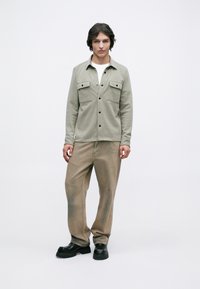 Νεαρός άνδρας που στέκεται φορώντας μπεζ πουκάμισο με κουμπωτές τσέπες, λευκό πουκάμισο, φαρδιά καφέ παντελόνια και μαύρα ογκώδη παπούτσια σε απλό φόντο.