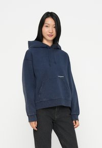 Marineblå kort hoodie med frontlomme, snørehette og en liten logo, kombinert med svarte jeans, laget av mykt materiale.