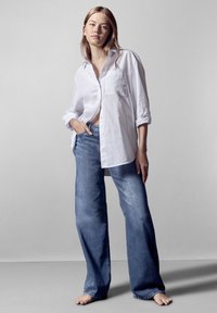Camisa blanca de gran tamaño con botones y un bolsillo, combinada con pantalones vaqueros holgados de mezclilla azul. Tejido suave con mangas con puños y diseño de pierna ancha.