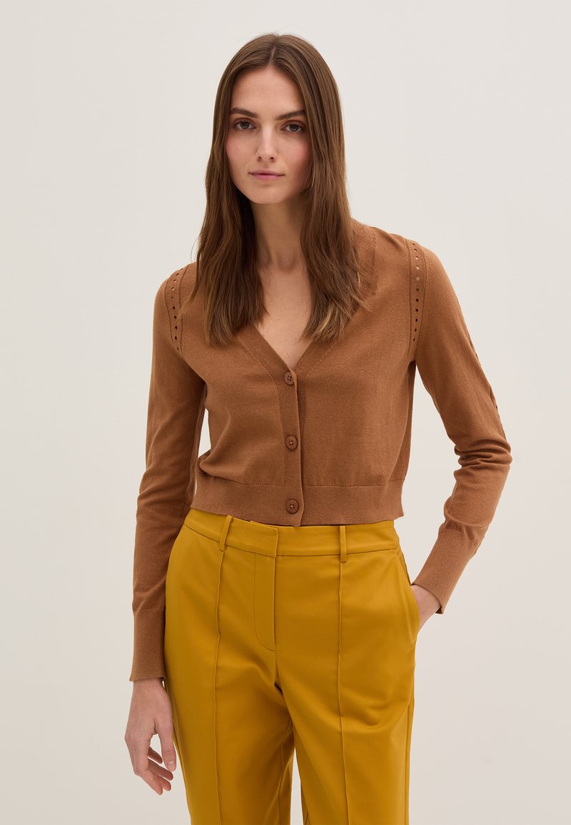 Brauner gestrickter Cardigan mit tiefem V-Ausschnitt, fünf vorderen Knöpfen und dekorativem Stickmuster entlang der Ärmel, getragen über gelben Hosen.