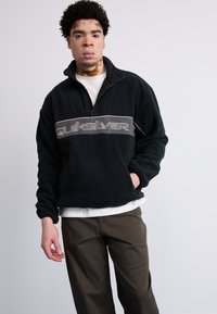 Czarny polarowy sweter z ćwierć zamkiem i szarym logo "Quiksilver" na piersi. Posiada luźny krój i boczne kieszenie.