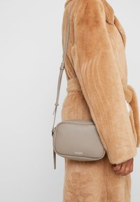 Malá béžová kožená crossbody taška s texturovaným povrchem, vybavená zipovým uzavírán�ím a nastavitelným popruhem. Výrazně zobrazené logo značky.