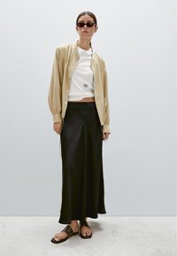 Veste beige clair à boutons sur une chemise blanche courte, associée à une jupe satin noire et des sandales plates noires. Design minimaliste, textures lisses.