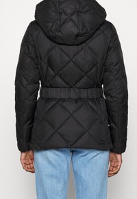 Sort quiltet jakke med hætte, bælte i taljen og diamantmønster. Glat tekstur og en afslappet pasform, kombineret med lyseblå jeans.