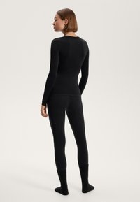 Zwart aangesloten languit-sleeve top en legging, gemaakt van een gladde stof. Model draagt dikke gebreide zwarte sokken, geposeerd van achteren tegen een neutrale achtergrond.