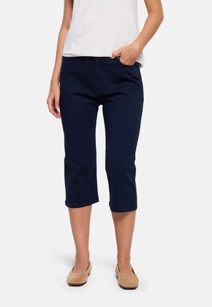 Mujer con pantalones azul marino cortos, camiseta blanca y zapatos planos beige, de pie con una mano en el bolsillo frente a un fondo liso.