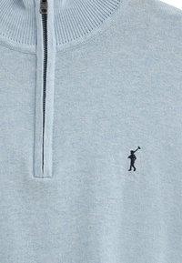 Ljust blå zip-up tröja med en ribbad krage och en liten svart broderad figur på vänster bröst. Mjuk stickad textur.