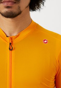Maillot de cyclisme orange avec un motif texturé, col court, fermeture éclair sur le devant et un logo rouge sur l'épaule gauche.