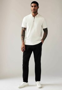 Valkoinen lyhythihainen polo, jossa on vetoketjukaulus, yhdistettynä mustiin slim fit -housuihin ja valkoisiin tennareihin. Tatuoinnit näkyvät käsissä.