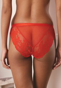 Rote Unterwäsche aus durchsichtiger Mesh und Spitzenverzierungen. Das Design verfügt über einen hoch ausschneidenden Rücken und florale Spitzenakzente an den Seiten.