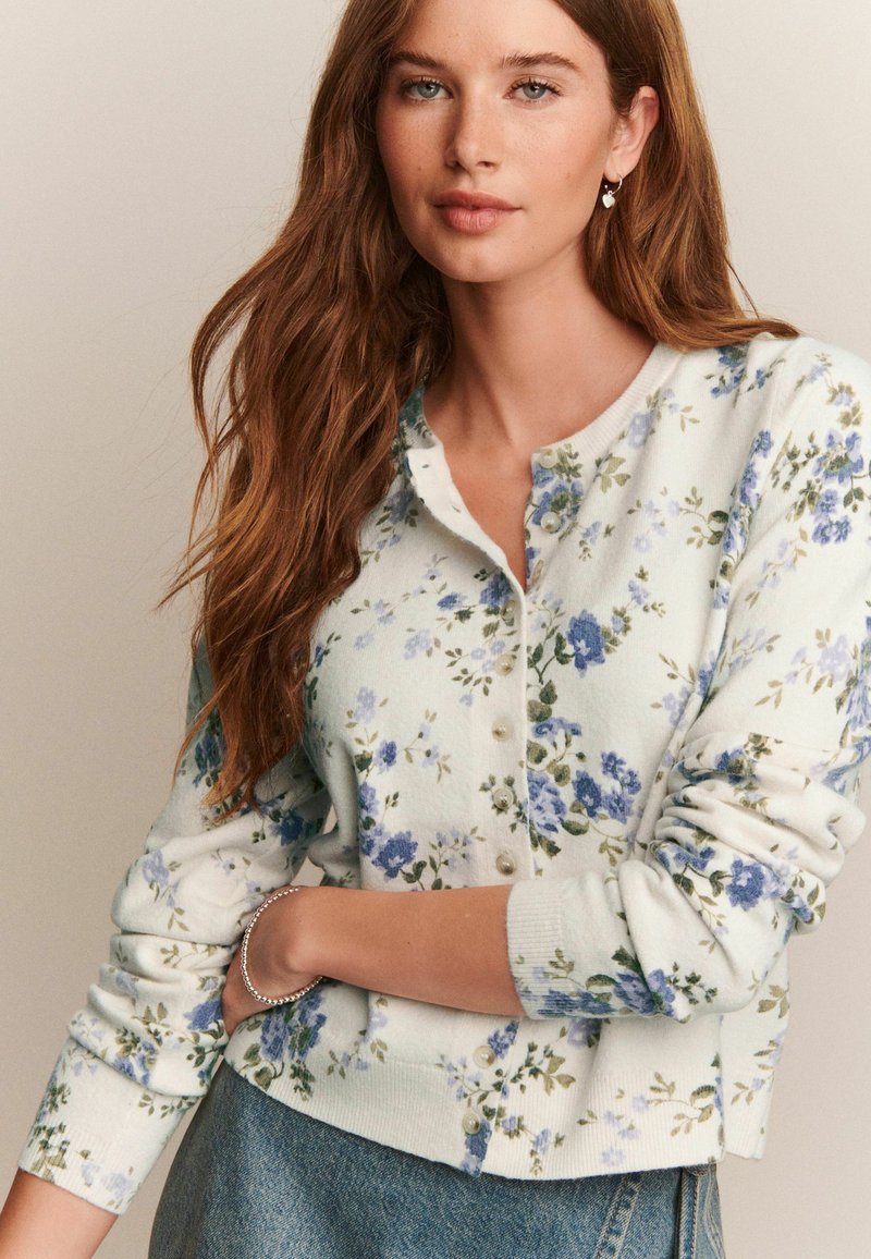 Femme aux longs cheveux bruns portant un cardigan fleuri blanc à boutons avec des fleurs bleues et un jean, posant les bras croisés.