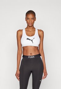 Hvit sport-bh med svart Puma-logo, med brede stropper og glatt stoff. Kombinert med svarte leggings som har "PUMA" på midjen.