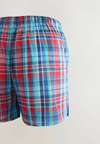 Sous-vêtements boxers amples pour hommes avec ceinture élastique et motif à carreaux rouges, bleus, verts et blancs sur fond clair.