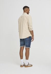 Chemise beige clair à manches longues, short en denim avec ourlets repliés, chaussettes blanches et baskets marron clair, vues de dos.