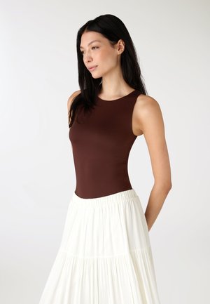 Donna con capelli lunghi e scuri che indossa un top marrone senza maniche e una gonna bianca a balze, in piedi davanti a uno sfondo semplice.