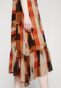 Main reposant contre une jupe midi plissée multicolore avec des rayures verticales abstraites dans des tons marron, orange, beige et noir.