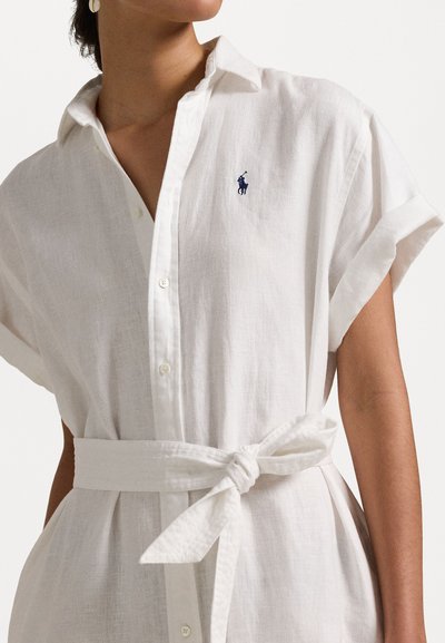 Robe chemise en lin blanc avec des manches courtes à revers, patte de boutonnage à l'avant et une ceinture en tissu nouée à la taille. Présente un petit logo bleu sur la poitrine.
