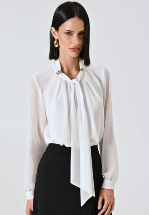Blusa di seta bianca con fiocco drappeggiato al collo, arricchita da dettagli in metallo dorato. Maniche lunghe e silhouette fluida. Abbinata a una gonna nera.