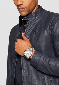 Marine læderjakke med syet detaljering, lynlåslukning og tryknapkrave; sølvur med tekstureret urskive og metalarmbånd på højre håndled.