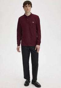 Polo a maniche lunghe borgogna con pattina dei bottoni e logo ricamato, abbinato a pantaloni neri e scarpe nere. Design semplice, forma aderente.