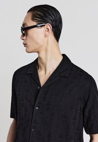 Camicia nera a maniche corte con un motivo geometrico a triangolo, caratterizzata da un design button-down e una texture morbida. Indossata con occhiali da sole scuri.