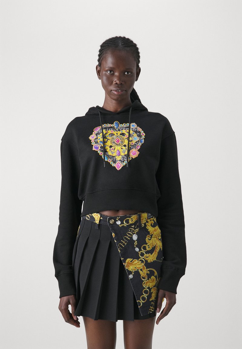 Haut de sweat noir court avec un design en forme de cœur coloré ; associé à une jupe plissée noire ornée de motifs baroques jaunes et d'accents en argent.