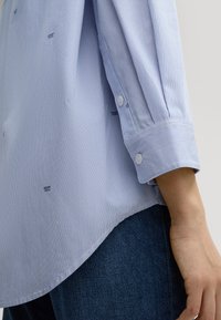 Hellblau gestreiftes Hemd mit geknöpften Manschetten, das einen leicht abgerundeten Saum und ein dezentes Logo in Dunkelblau nahe dem Saum aufweist. Kombiniert mit dunklem Denim.