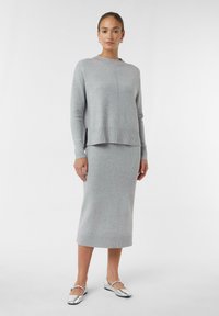 Szary dzianinowy sweter z okrągłym dekoltem i bocznymi rozcięciami, zestawiony z pasującą, długą szarością spódnicą. Modelka nosi srebrne płaskie buty.