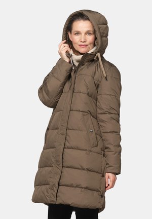 Woman wearing a brown hooded puffer coat, holding the hood with one hand, standing against a plain light background.