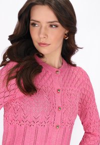 Jeune femme aux longs cheveux bruns portant des boucles d'oreilles dorées et un cardigan en tricot rose texturé avec des boutons métalliques, regardant vers sa gauche.