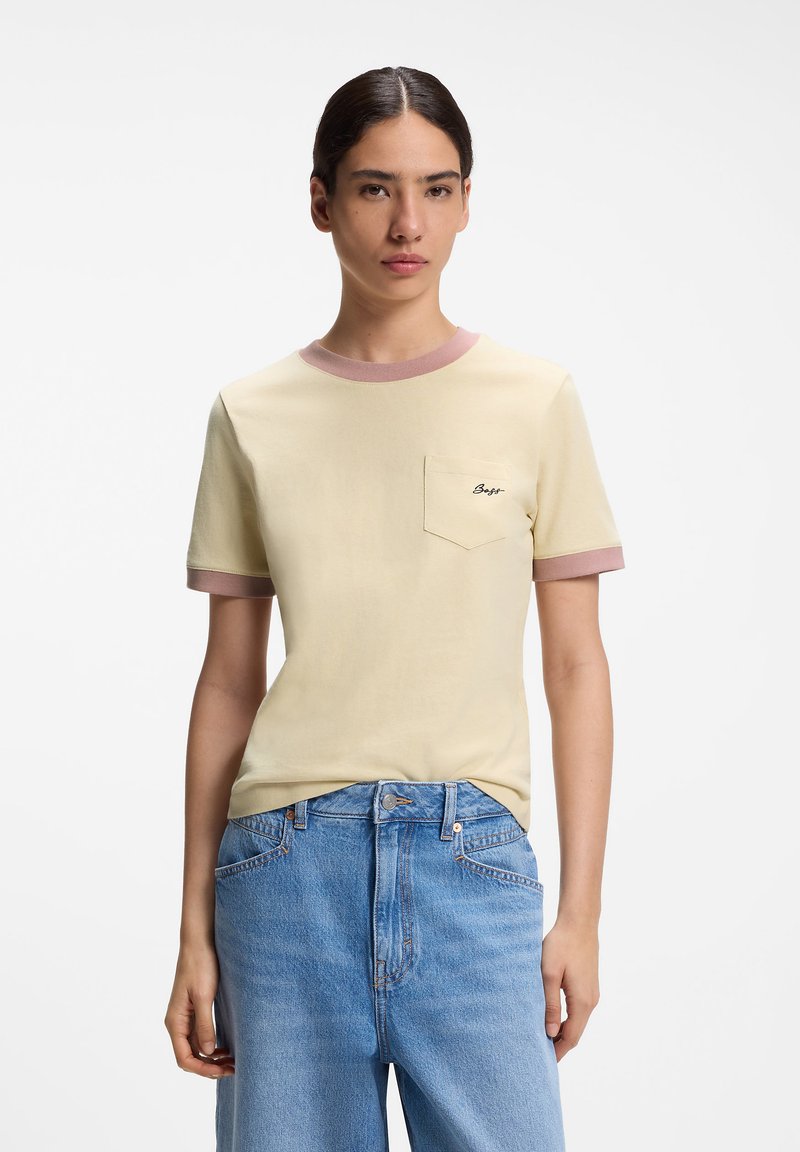 Νέος με σκούρα μαλλιά που φοράει ένα κρεμ T-shirt με ροζ λεπτομέρειες και μικρό λογότυπο στην τσέπη, σε συνδυασμό με ανοιχτό μπλε τζιν με ψηλή μέση.