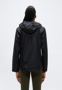 Άτομο που φοράει μαύρο μπουφάν με κουκούλα και χακί παντελόνι, στέκεται με την πλάτη προς το μέρος μας σε ένα απλό φωτεινό φόντο.