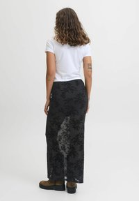 Gonna lunga nera trasparente con motivo in pizzo floreale, abbinata a una t-shirt bianca aderente. Presenta uno spacco alto e stivali marroni alla caviglia.