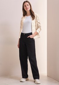 Chaqueta beige claro con cremallera y textura estampada, camisa blanca ajustada, pantalones de pierna ancha negros y zapatillas blancas. Calzado plano mostrado.