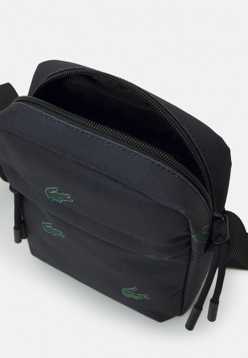 Sac bandoulière noir en tissu avec fermeture éclair, orné d'accents verts en logo crocodile. Design compact avec une surface texturée et une sangle ajustable.