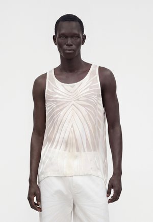 Modèle masculin à la peau foncée portant un débardeur blanc transparent avec un motif géométrique et un pantalon blanc, debout devant un fond uni.