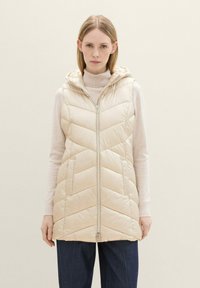 Padded, hellbeige Weste mit Kapuze, die einen Reißverschluss und ein Zickzack-Steppmuster aufweist. Getragen über einem cremefarbenen Pullover und dunklen Hosen.