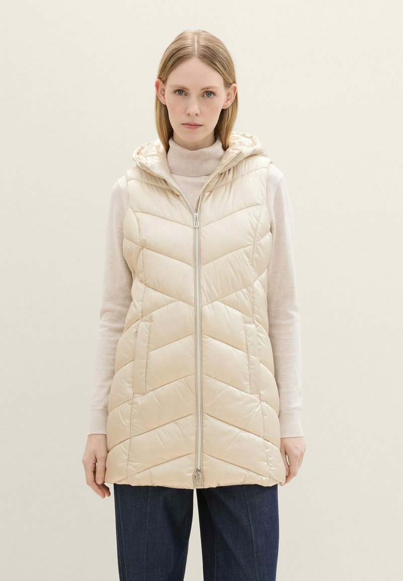 Padded, hellbeige Weste mit Kapuze, die einen Reißverschluss und ein Zickzack-Steppmuster aufweist. Getragen über einem cremefarbenen Pullover und dunklen Hosen.