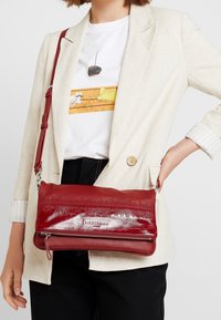 Červená kožená kabelka přes rameno s texturovaným povrchem a lesklým povrchem; obsahuje zipový uzávěr a nastavitelný popruh.