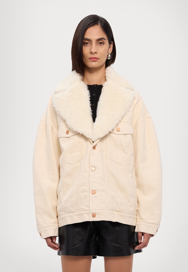 Chaqueta de mezclilla beige oversize con cuello de piel sintética, que cuenta con grandes bolsillos y botones de cobre. Textura suave, corte relajado.