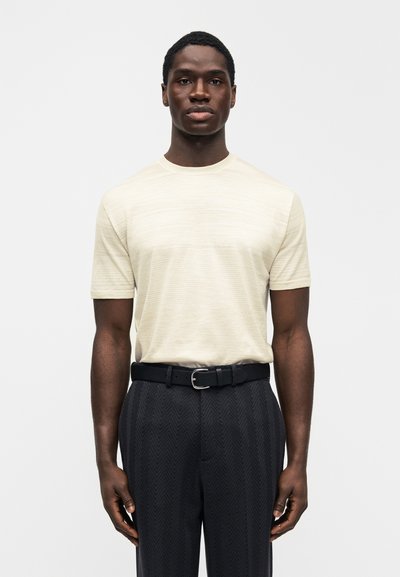 Ανοιχτό μπεζ πουκάμισο με κοντά μανίκια και διακριτική ραβδωτή υφή, συνδυασμένο με σκούρο παντελόνι με σχέδια και μαύρη ζώνη, εστιάζοντας στη γραμμή και την απλότητα.