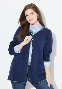Jonge vrouw met lang bruin haar, gekleed in een blauwe cardigan over een geruite blouse en jeans, terwijl ze een knoop aan de cardigan vastmaakt.