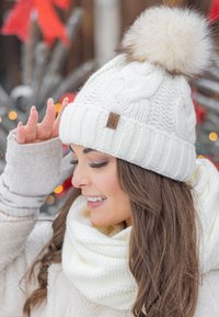 Hvid strikket hue med en fluffy pompon, præget af et vævet kabeldesign og et brunt mærke. Begge dele ledsages af et lysefarvet tørklæde.
