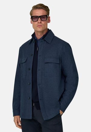 Homme portant une veste boutonnée bleu foncé, un pantalon foncé et des lunettes de soleil noires, debout avec une main dans la poche sur fond blanc.