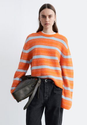 Jeune femme portant un pull à rayures orange et bleu clair, pantalon noir, tenant un sac à main en cuir noir sur un fond uni.