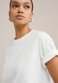T-shirt blanc en coton avec des manches retroussées et un col rond, présentant une texture lisse et un design simple. Aucun motif ni accent visible.