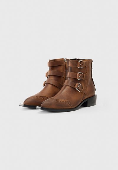 Botas de tobillo de cuero marrón con tres correas ajustables, hebillas de plata, puntas afiladas, perforaciones decorativas y un tacón bajo tipo bloque.